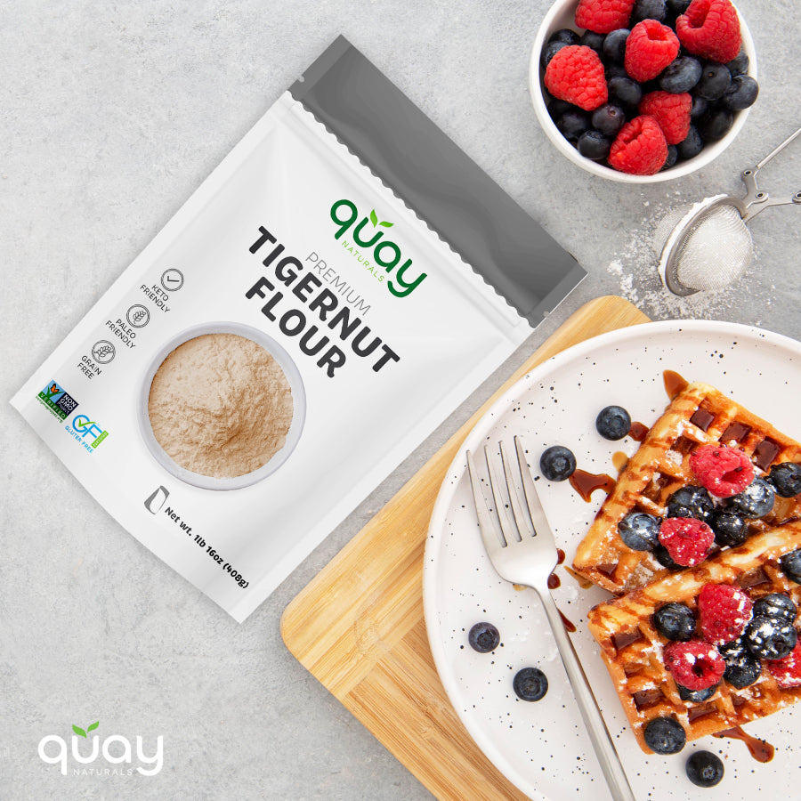 A white plate with two waffles with berries and powdered sugar, alongside a fork on a wooden board & with A bag of Quay Naturals Premium Tigernut Flour and a bowl of berries.